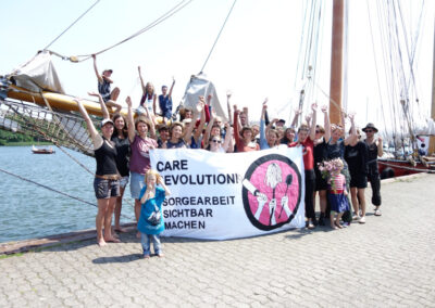 Eine Gruppe Menschen steht vor einem Segelboot und hält jubelnd die Arme in die Luft. Auf dem Banner, das sie vir sich halten, steht "Care Revolution Sorgearbeit sichtbar machen"