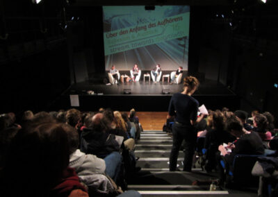 Ein Publikum blickt auf eine Bühne mit vier Diskussionteilnehmenden. Hinter der Bühne ist eine Leinwand, auf der "Über den Anfang des Aufhörens, Reflexion & Austausch stream towards degrowth" steht