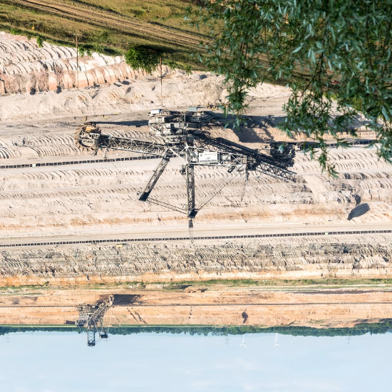 ein umgedrehtes Bild von einem Braunkohletagebau. Der blaue Himmel ist im unteren Teil des Bildes. Im oberen Teil sieht eins grüne Zweige und Fläche. Mittig ist ein Kohle-Bagger zu sehen, der Abraum abbaggert.
