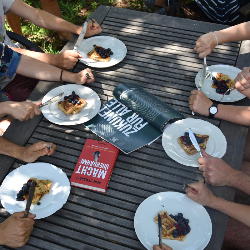 Sechs Menschen essen gemeinsam an einem Tisch und essen French Toast, in der Mitte das Buch "Machtübernahme" von Arne Semsrott