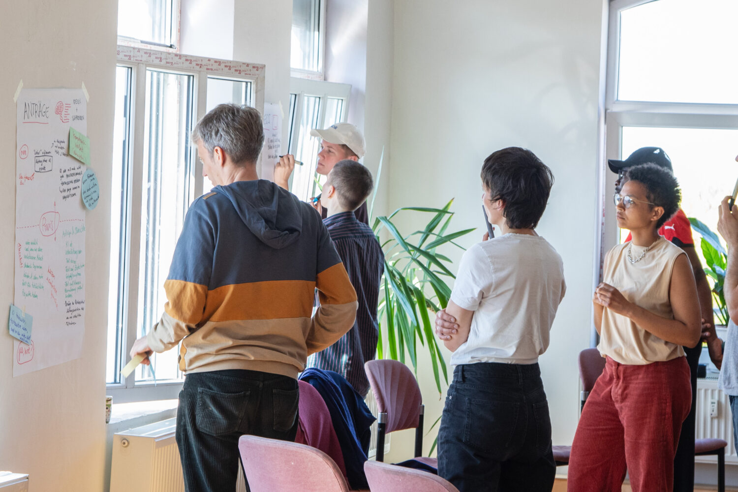 Eine Gruppe von vier Personen steht vor einem Fenster und betrachtet ein Plakat an der Wand. Eine Person trägt einen gestreiften Pullover, eine andere ein weißes T-Shirt. Im Hintergrund sind Pflanzen und Stühle sichtbar.